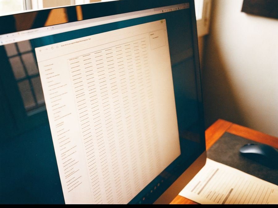 Un monitor de oficina visto de lado, con un portal web lleno de filas de una tabla, y al lado el rat&oacute;n y un impreso en papel.